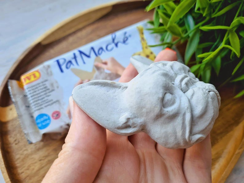 Pode incluir: Uma cabe&ccedil;a de bulldogue franc&ecirc;s esculpida em cinza, feita de papel mach&eacute;. A cabe&ccedil;a est&aacute; sendo segurada em uma m&atilde;o, e o fundo mostra um pacote de papel mach&eacute;.