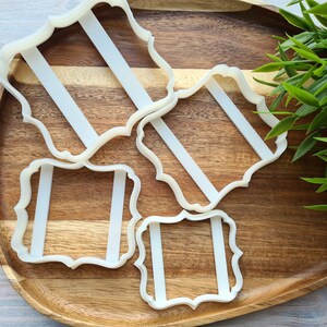 May include: Four white plastic cookie cutters in the shape of ornate frames on a wooden surface. The cutters are arranged in a staggered pattern.