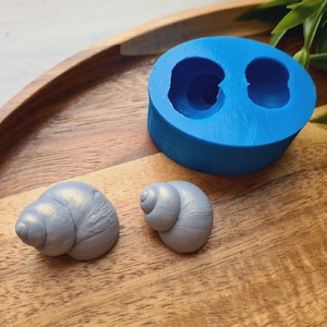 Pode incluir: Um molde de silicone azul brilhante com duas cavidades circulares ao lado de duas formas de concha de caracol acabadas em um tom prateado metálico. O molde apresenta um exterior liso e arredondado com impressões profundas para moldar espirais.
