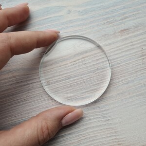 May include: A clear round glass disc, approximately 2 inches in diameter, on a light wood surface.