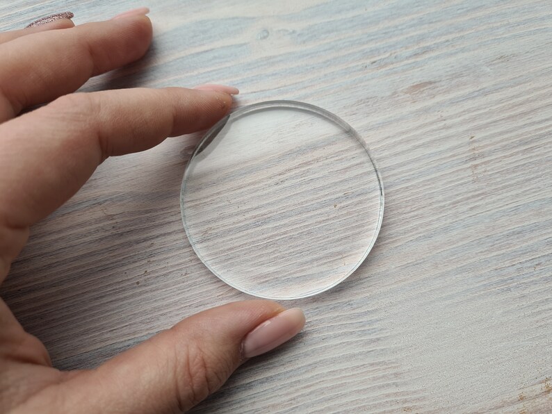 May include: A clear round glass disc, approximately 2 inches in diameter, on a light wood surface.