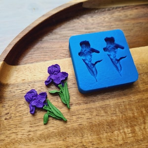 Peut inclure: Deux fleurs d'iris violettes faites à la main avec des tiges et des feuilles vertes sont posées à côté d'un moule en silicone bleu avec deux empreintes de fleurs d'iris. Les articles sont sur un plateau en bois.