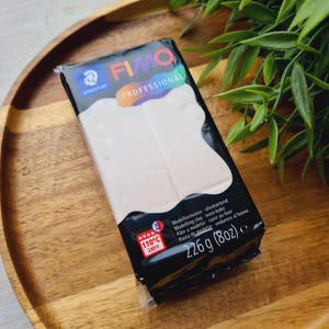 May include: A package of white FIMO Professional polymer clay, 226 g (8oz), on a wooden tray. The black and white packaging includes the brand name, product details, and oven baking instructions at 230°F (110°C). Green leaves are in the background.