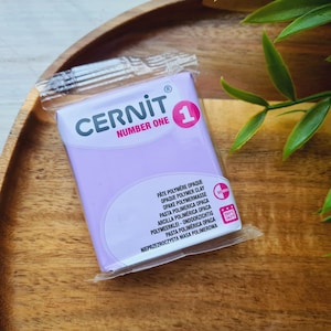 May include: A package of Cernit Number One polymer clay in a light purple color. The package is sealed in clear plastic and rests on a wooden tray. The text on the package includes the brand name and product details.