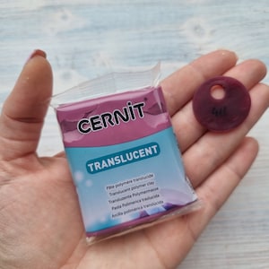 May include: A package of translucent polymer clay in a pink color. The package has the brand name "CERNIT" and the text "TRANSLUCENT" printed on it. The package is next to a small, round, pink clay disc with a hole in the center and the number "48" printed on it.