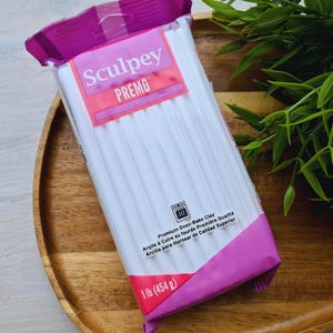 Op de afbeelding: Een verpakking witte Sculpey Premo oven-bakklei. De verpakking is roze en paars en vermeldt "Premium Oven-Bakklei" en "1 lb (454 g)".