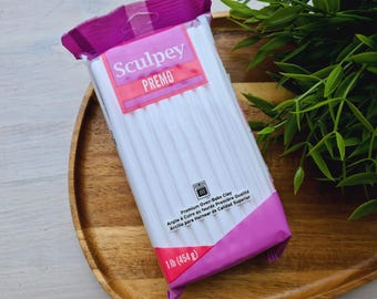 Puede incluir: Un paquete de arcilla para hornear Sculpey Premo blanca. El paquete es rosa y morado y dice "Arcilla para hornear de calidad superior" y "1 lb (454 g)".