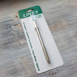 Puede incluir: Un cuchillo de arte plateado con un embalaje verde y blanco. El embalaje dice "Art Knife / Design Cutter / Couteau arts graphiques / cuchilla arte" y "LINEX".