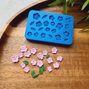 May include: A blue silicone mold with flower and leaf shapes sits on a wooden tray. Scattered around are small, pink flower and green leaf-shaped decorations, likely for crafting or baking. The scene is well-lit.
