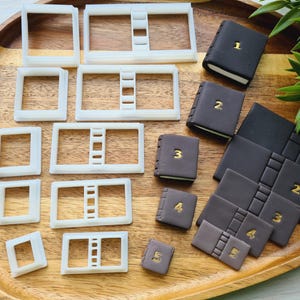 Pode incluir: Um conjunto de 10 cortadores de biscoitos de plástico branco em forma de livros e 5 livros de fondant marrom com números dourados de 1 a 5. Os cortadores de biscoitos estão dispostos em filas sobre uma superfície de madeira.