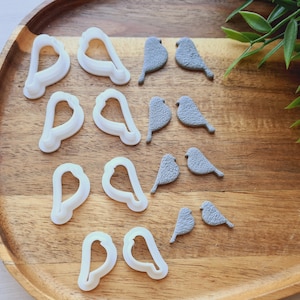 May include: A wooden tray holds white bird-shaped cookie cutters and gray bird-shaped clay ornaments. The cookie cutters are in the shape of a bird's body. The clay birds have textured details.