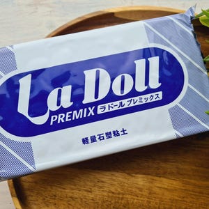May include: A package of La Doll Premix clay, featuring a blue and white design with Japanese text. The package is rectangular and rests on a wooden surface, with green foliage in the background. The text "La Doll" is prominently displayed.