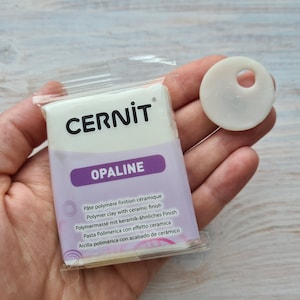 Może przedstawiać: Opakowanie białej gliny polimerowej z ceramicznym wykończeniem. Opakowanie jest oznaczone "CERNIT OPALINE" i zawiera instrukcje w wielu językach. Obok opakowania pokazana jest mała próbka gliny.