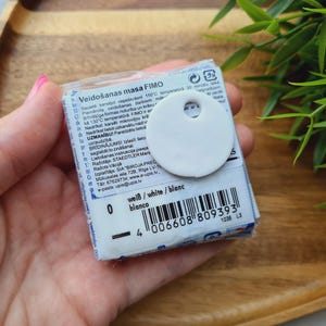 May include: A package of white FIMO modeling clay with a white circular tag attached. The package has text in multiple languages, including "wei&szlig;/white/blanc" and a barcode. The clay is held in a hand.