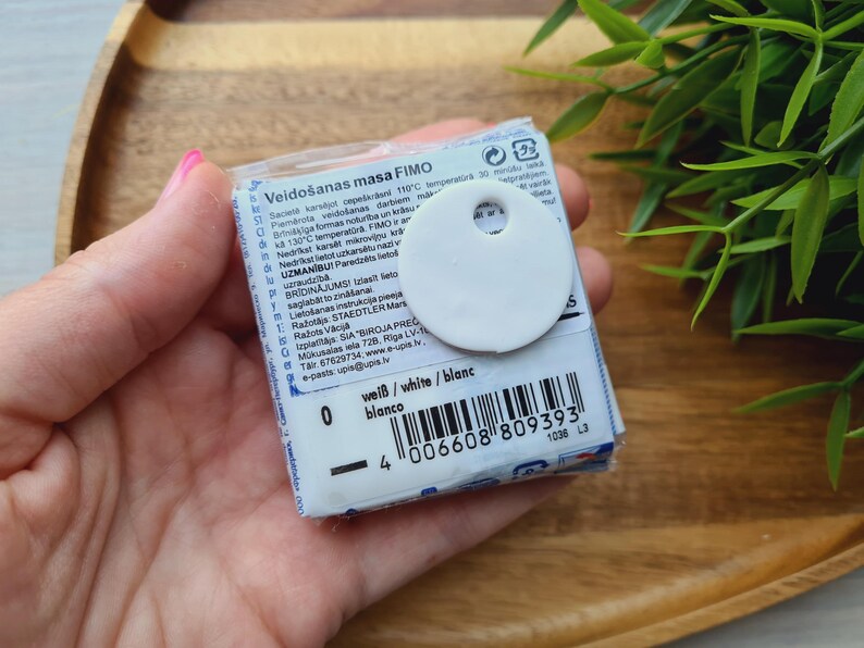 May include: A package of white FIMO modeling clay with a white circular tag attached. The package has text in multiple languages, including "wei&szlig;/white/blanc" and a barcode. The clay is held in a hand.