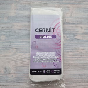 Może przedstawiać: Opakowanie gliny polimerowej Cernit Opaline, 500g, z ceramicznym wykończeniem. Białe opakowanie ma fioletową etykietę z nazwą marki i szczegółami produktu. Dołączono instrukcje pieczenia.