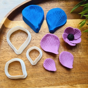 Puede incluir: Un juego de cortadores de galletas con forma de flor en varios tamaños, que incluye un pétalo azul grande, un pétalo azul más pequeño, un pétalo morado, un pétalo morado más pequeño, un círculo blanco pequeño, un óvalo blanco pequeño y una forma de lágrima blanca pequeña. Los cortadores están sobre una superficie de madera.