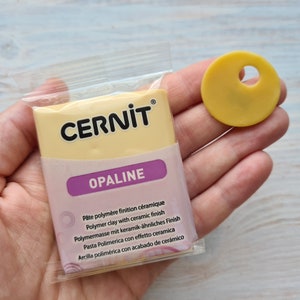 Może przedstawiać: Opakowanie żółtej gliny polimerowej z etykietą "CERNIT OPALINE" i opisem w kilku językach. Opakowanie znajduje się w przezroczystym plastikowym woreczku. Obok opakowania znajduje się mały żółty dysk z gliny z otworem w środku.