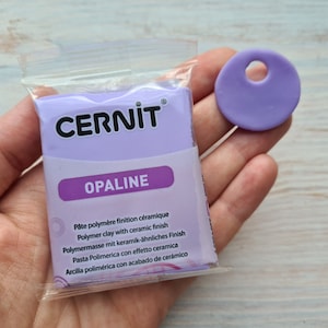 Może przedstawiać: Opakowanie fioletowej gliny polimerowej z etykietą "CERNIT OPALINE" i opisem gliny w wielu językach. W tle widać mały fioletowy krążek z gliny z otworem w środku.