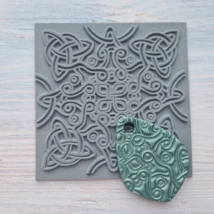 Pode incluir: Um carimbo de silicone cinza com um design de nó celta. Uma impressão verde-azulada do carimbo é mostrada ao lado.