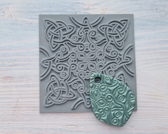 Pode incluir: Um carimbo de silicone cinza com um design de nó celta. Uma impressão verde-azulada do carimbo é mostrada ao lado.