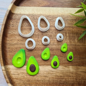 Puede incluir: Un juego de cortadores de galletas de plástico blanco en forma de aguacate y formas de aguacate de fondant verde y marrón sobre una superficie de madera.