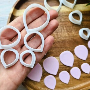 Może przedstawiać: Zestaw białych plastikowych foremek do ciasteczek w kształcie płatk&oacute;w kwiat&oacute;w, ułożonych na drewnianej powierzchni z kilkoma r&oacute;żowymi płatkami kwiat&oacute;w z lukru.