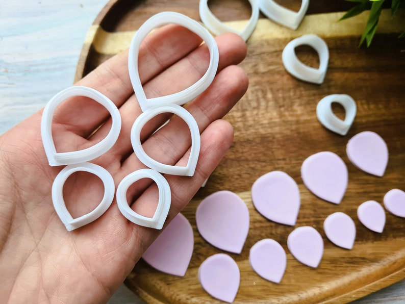 Może przedstawiać: Zestaw białych plastikowych foremek do ciasteczek w kształcie płatk&oacute;w kwiat&oacute;w, ułożonych na drewnianej powierzchni z kilkoma r&oacute;żowymi płatkami kwiat&oacute;w z lukru.
