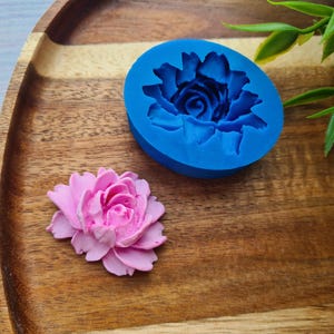 May include: A blue silicone mould for making a pink flower shaped cake decoration. The mould is on a wooden surface.