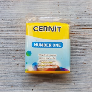 May include: A yellow package of CERNIT Number One polymer clay. The package has text in multiple languages, including "Pâte polymère opaque", "Opaque polymer clay", "Opake Polymermasse", "Pasta Polimerica opaca", and "Arcilla polimerica opaco".