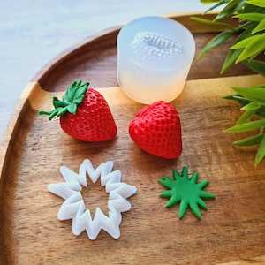 Pode incluir: Um conjunto de moldes de silicone para fazer guloseimas com formato de morango. Os moldes estão sobre uma superfície de madeira com dois morangos vermelhos e um molde em forma de estrela branco e verde.
