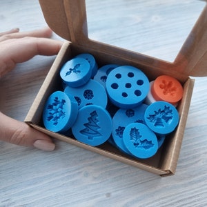 May include: A box of blue silicone molds for baking, featuring various holiday-themed designs, including snowflakes, holly leaves, and Christmas trees. The box also includes a red snowflake mold.