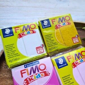 May include: Four packages of Fimo Kids oven-bake modeling clay. The packages are yellow, white, pink, and light blue. Each package has the Fimo Kids logo and instructions for baking the clay at 110 degrees Celsius or 230 degrees Fahrenheit.