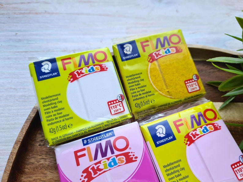 May include: Four packages of Fimo Kids oven-bake modeling clay. The packages are yellow, white, pink, and light blue. Each package has the Fimo Kids logo and instructions for baking the clay at 110 degrees Celsius or 230 degrees Fahrenheit.