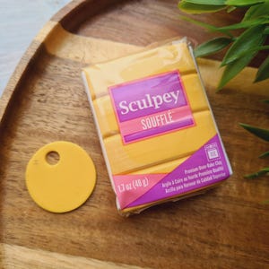 Puede incluir: Un paquete de arcilla polimérica Sculpey Souffle amarilla, con una etiqueta amarilla a juego. El paquete es de 48 g. El texto en el paquete dice "Sculpey Souffle". Los artículos están en una bandeja de madera.