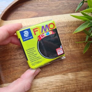 May include: A black FIMO Kids oven-bake modelling clay in a 1.5 ounce package. The package has a yellow background with the FIMO Kids logo and the text "Modelliernusse ofenh&auml;rtend Modelling clay oven-bake P&acirc;te &agrave; modeler cuire au four Pasta de modelar endurence al horno 42g (1.5oz) e MADE IN GERMANY FABRIQUE EN ALLEMAGNE".