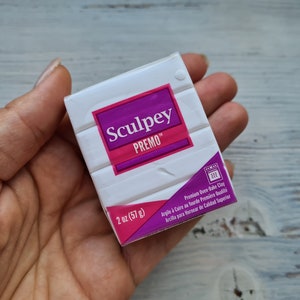 May include: A hand holds a package of white Sculpey Premo oven-bake clay. The package features a purple and pink label with the brand name and product details. It contains 57 grams of clay.