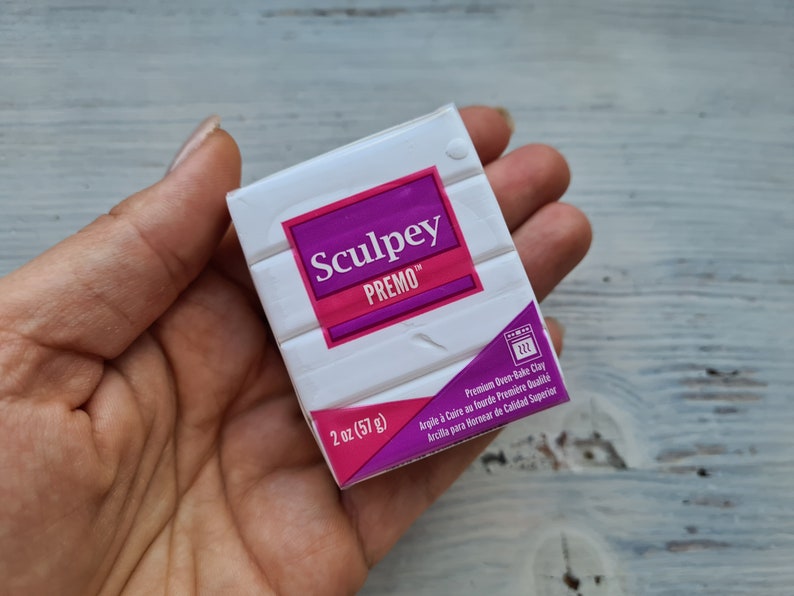 May include: A hand holds a package of white Sculpey Premo oven-bake clay. The package features a purple and pink label with the brand name and product details. It contains 57 grams of clay.