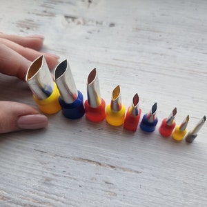 Puede incluir: Una colección de terminales de crimpado plateados con fundas de plástico de colores. Las fundas son amarillas, azules y rojas. Los terminales están dispuestos en diagonal, siendo el más grande a la izquierda y disminuyendo de tamaño.