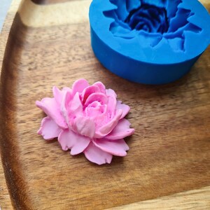 May include: A pink polymer clay flower, resting on a wooden surface alongside a blue silicone mould of a flower.