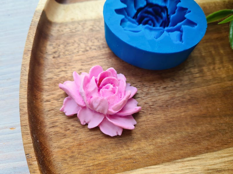 May include: A pink polymer clay flower, resting on a wooden surface alongside a blue silicone mould of a flower.