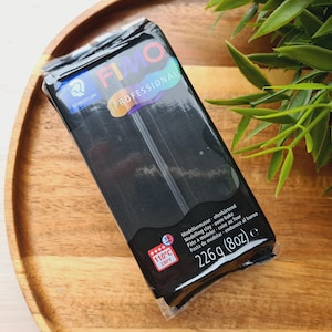 May include: A package of black FIMO Professional polymer clay, 226g (8oz), for oven-baking. The package has the brand name and product information printed on it. The package is on a wooden tray with green leaves in the background.