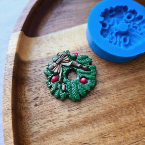 Puede incluir: Una corona de Navidad verde y marr&oacute;n con bayas rojas, un lazo marr&oacute;n y un molde de silicona azul en el fondo.