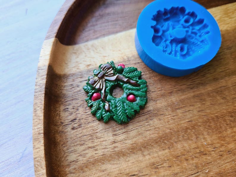 Puede incluir: Una corona de Navidad verde y marr&oacute;n con bayas rojas, un lazo marr&oacute;n y un molde de silicona azul en el fondo.