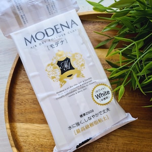 Könnte beinhalten: Eine Packung weißer lufttrocknender Polymer-Knete namens Modena. Die Verpackung hat ein goldenes Design mit dem Text "Modena is air dry clay soft & flexible. Dries with translucent & waterproof. Virtually unbreakable. Suitable for flowers, sweets & deco, doll." Die Verpackung enthält auch den Text "[ホワイト] White [半透明]" und "標準2509 水に強く、しなやかで丈夫 [最高級樹脂粘土]"