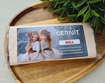 May include: A rectangular block of Cernit Doll modelling clay with a porcelain finish, wrapped in clear plastic. The label features an image of two children and the text Cernit Doll, Modelling clay with porcelain finish, Pâte à modeler finition porcelaine, Modelliermasse mit Porzellanschimmer.