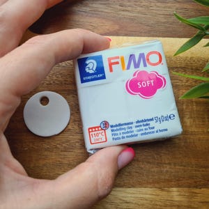 May include: A white package of FIMO SOFT modeling clay, 57g (2 oz), with a pink and white label. A small, round, white tag with a hole is next to the clay. The image is on a wooden surface.