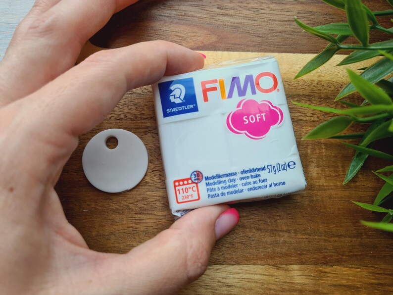 May include: A white package of FIMO SOFT modeling clay, 57g (2 oz), with a pink and white label. A small, round, white tag with a hole is next to the clay. The image is on a wooden surface.