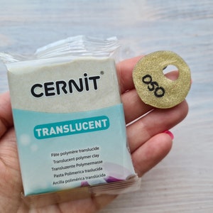 May include: A package of translucent polymer clay by the brand Cernit. The package is white with blue text and the brand name is in black. The clay is translucent and has a slight shimmer. The package is being held in a hand. A gold circle with the number 050 is also visible in the image.