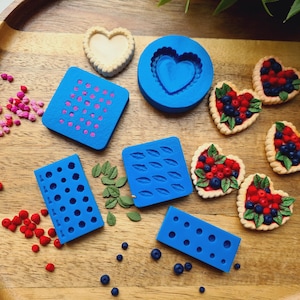 Peut inclure: Une collection de moules en silicone bleu pour créer des miniatures alimentaires, incluant des formes carrées, rectangulaires et circulaires. Plusieurs tartelettes aux baies en forme de cœur, des perles et des décorations sont éparpillées.